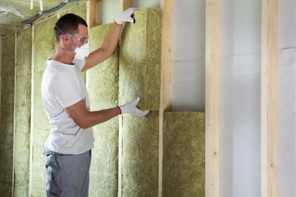 Worker in protective goggles and respirator insulating rock wool insulation in wooden frame for future house walls for cold barrier. Comfortable warm home, economy, construction and renovation concept